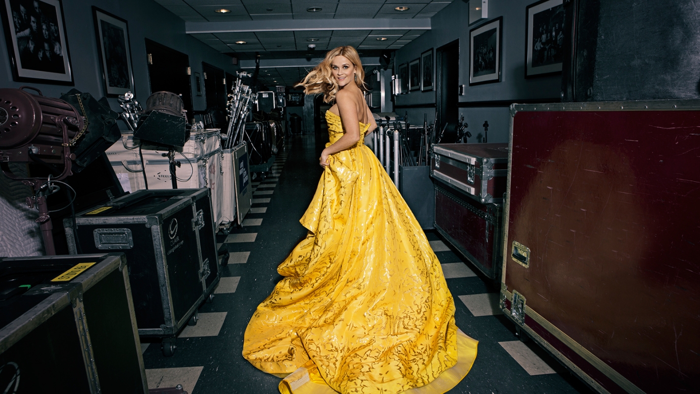 Blonde woman in a yellow gown turns her head in a hallway lined with road cases and lighting equipment, creating a captivating fashion image.