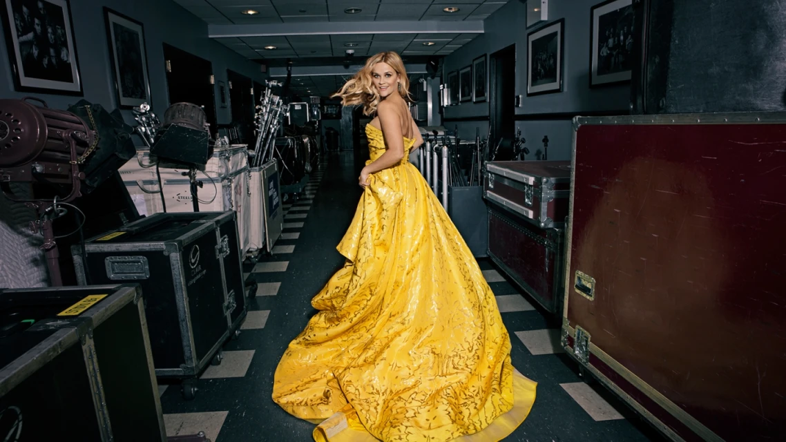 Blonde woman in a yellow gown turns her head in a hallway lined with road cases and lighting equipment, creating a captivating fashion image.