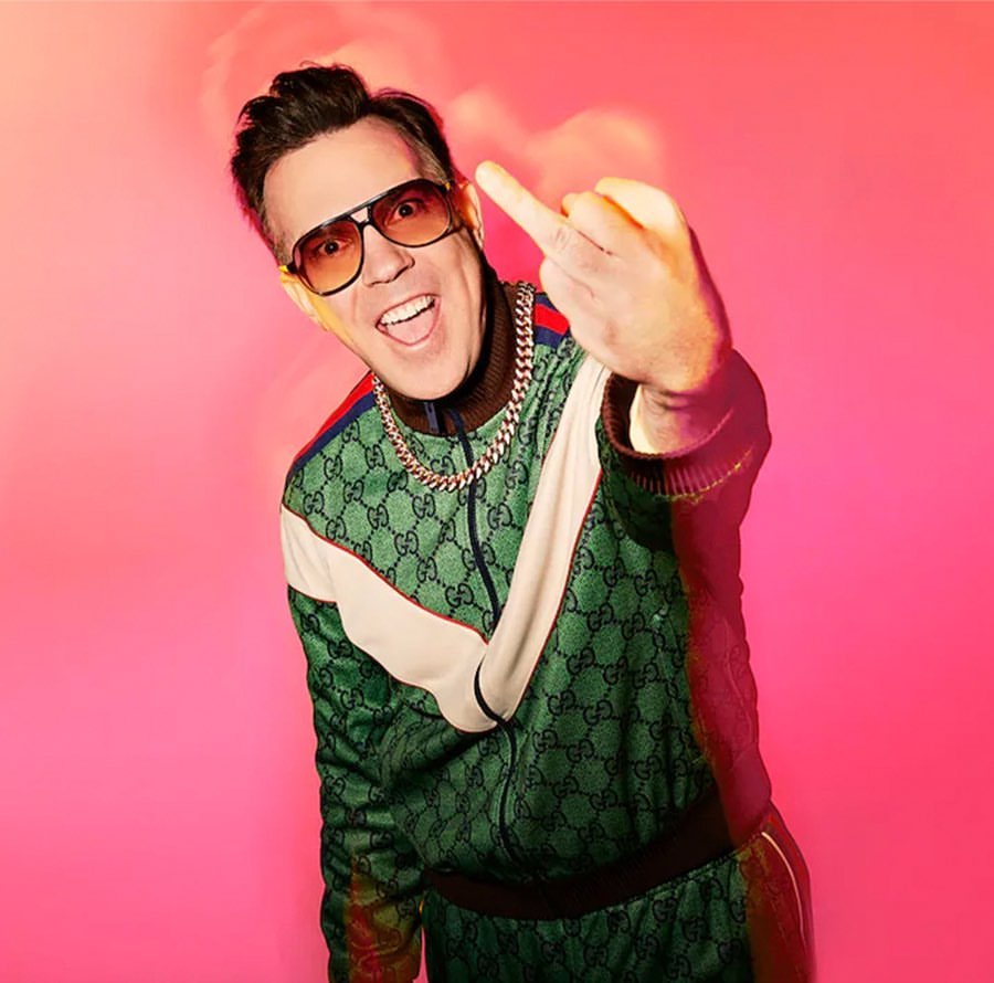 Adult male with a middle finger gesture wears sunglasses and a patterned green tracksuit in front of a bright pink backdrop.