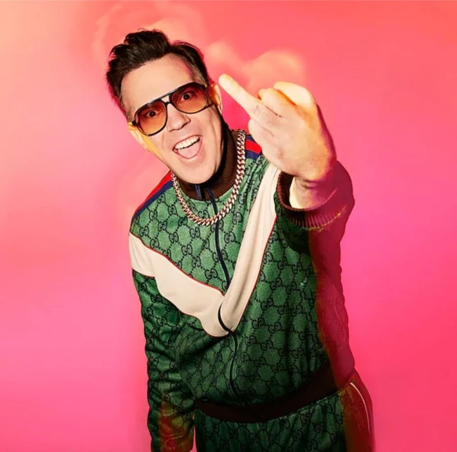 Adult male with a middle finger gesture wears sunglasses and a patterned green tracksuit in front of a bright pink backdrop.
