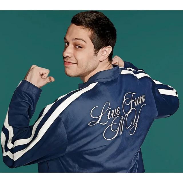 A man with short dark hair, fair skin, a navy blue jacket with white stripes, and embroidered text on the back poses indoors.