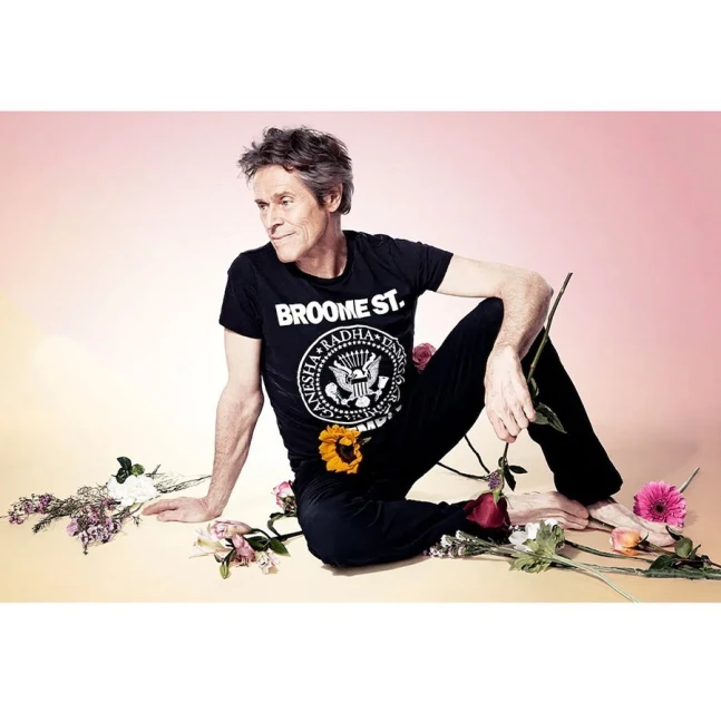 Portrait of a mature adult male sitting barefoot on the floor surrounded by various cut flowers and wearing a t-shirt and pants.