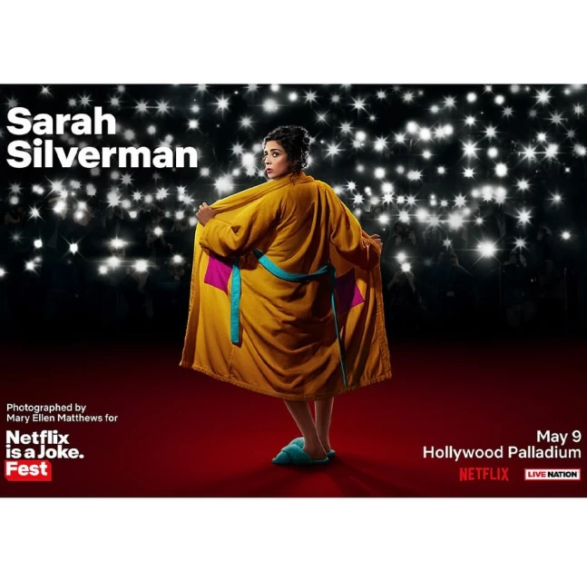Sarah Silverman in a yellow robe and slippers, standing on a red carpet in front of a background with many white lights.