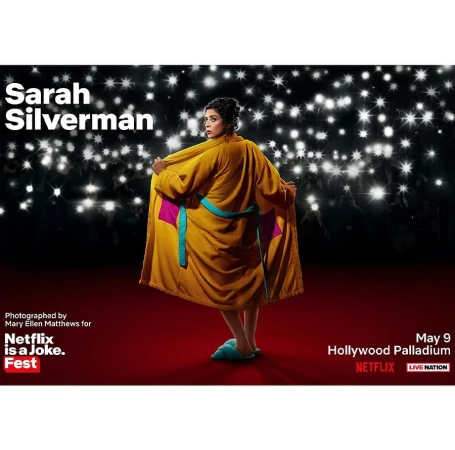 Sarah Silverman in a yellow robe and slippers, standing on a red carpet in front of a background with many white lights.