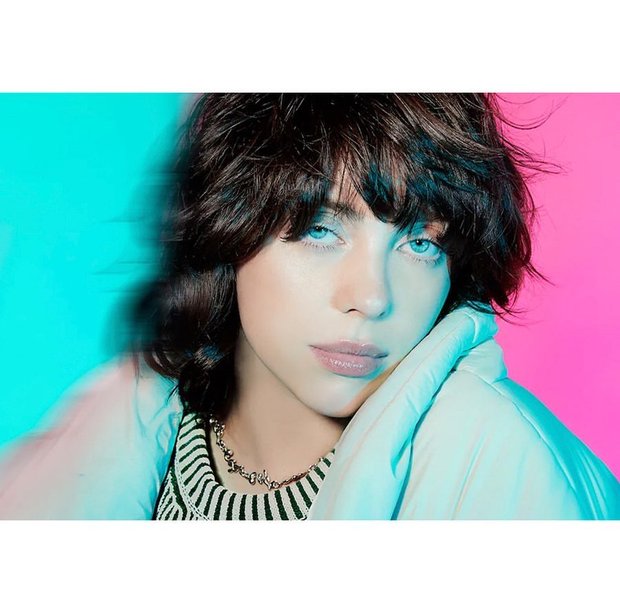 Close-up portrait of a young adult female with short, dark hair and light eyes, posing against a background split between cyan and pink.