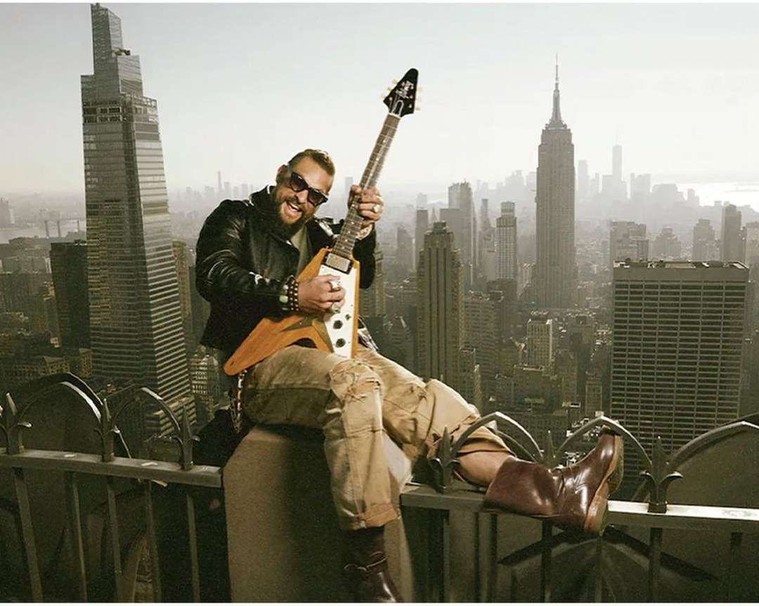 Adult male playing electric guitar atop building in New York City with skyline view including the Empire State Building.