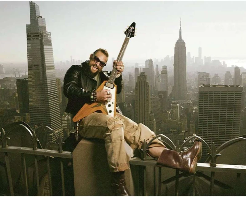 Adult male playing electric guitar atop building in New York City with skyline view including the Empire State Building.
