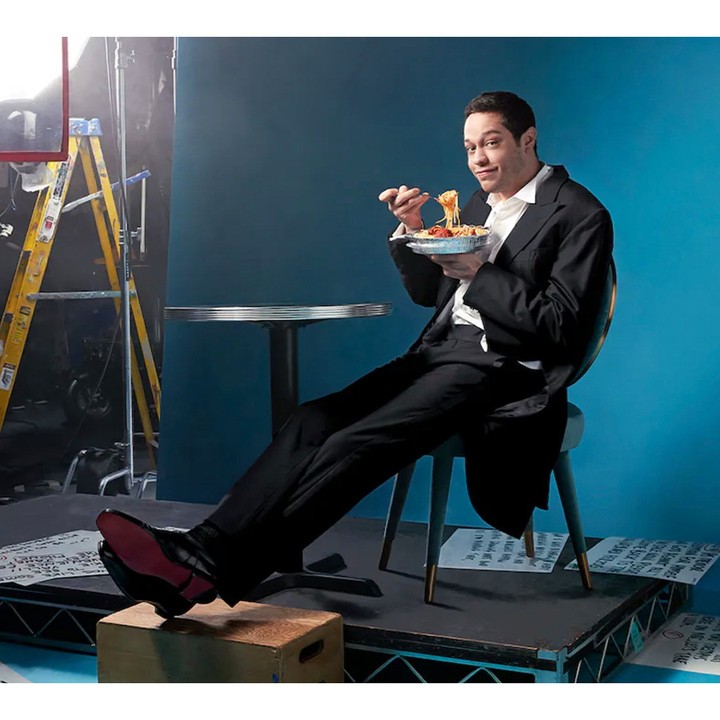 Pete Davidson eating spaghetti from a foil takeout container on a studio set, photographed by Mary Ellen Matthews.