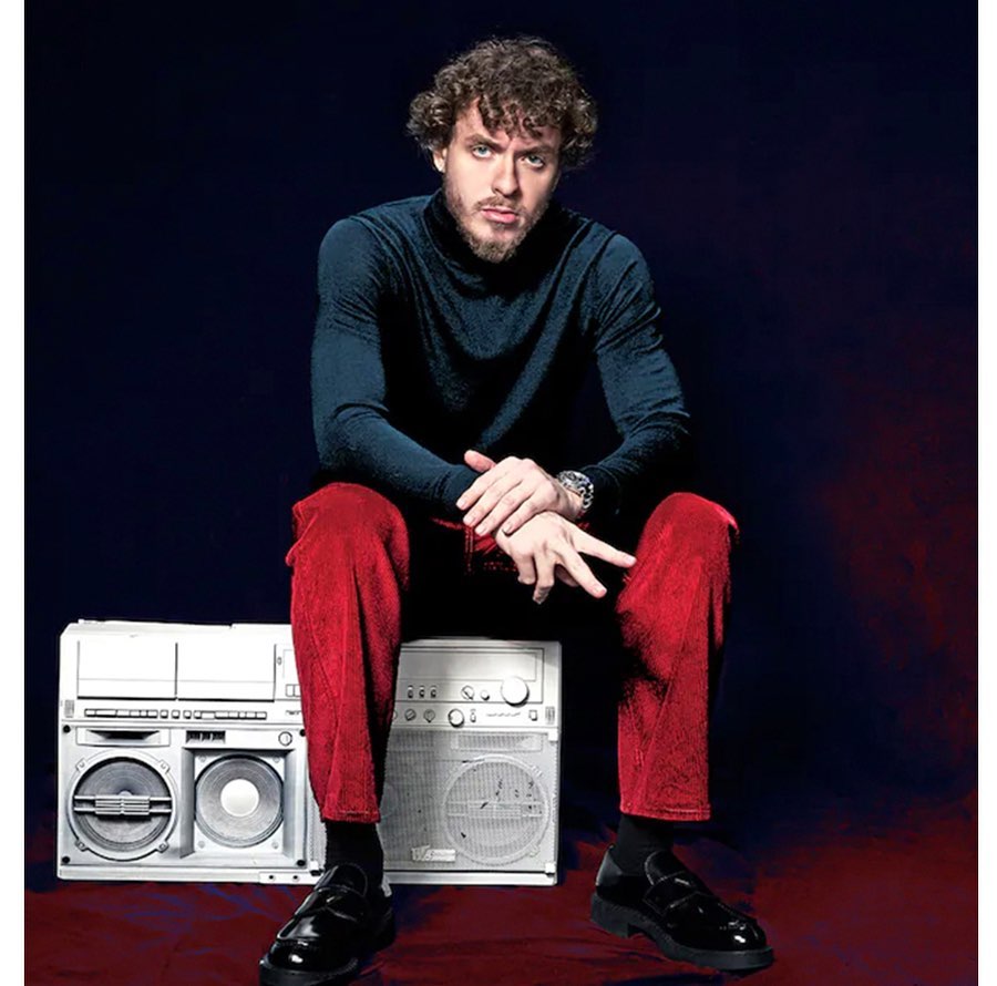 Portrait of a male model sitting on two vintage boomboxes in a studio wearing red corduroy pants, a dark blue turtleneck, and black loafers.