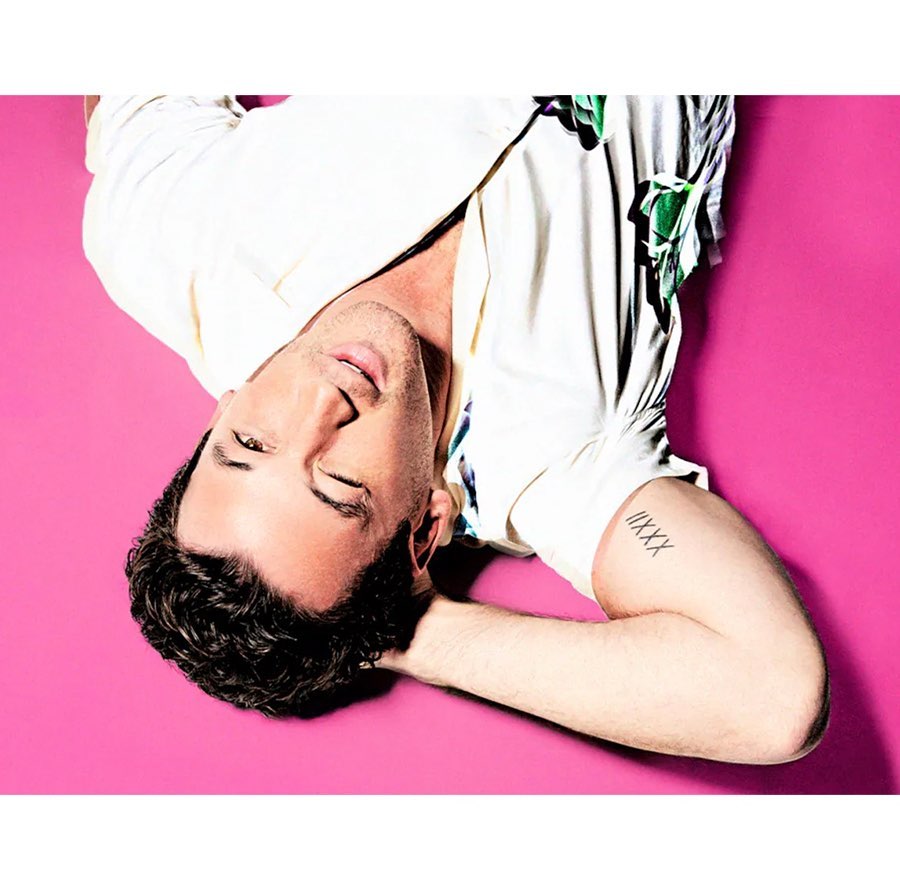 Portrait of a fair-skinned adult male laying down on a bright pink background, wearing a white floral shirt and featuring a visible arm tattoo.