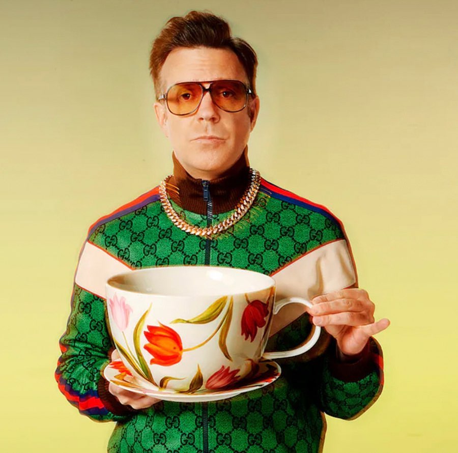 A man in a Gucci tracksuit and sunglasses holds a giant tea cup with floral design in a medium shot, set against a gradient yellow backdrop.