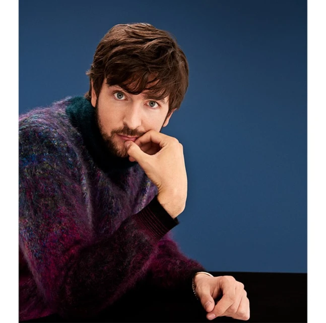 Portrait of a fair-skinned man with brown hair wearing a colorful mohair sweater against a blue background in a studio setting.