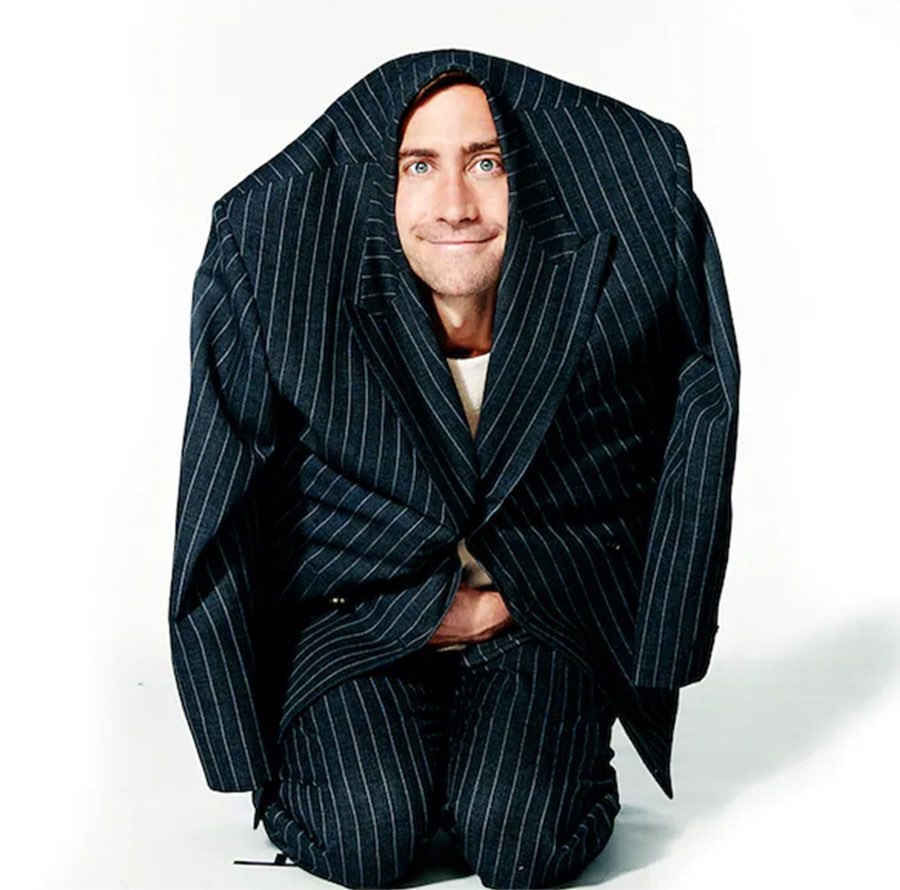 Smiling adult male with dark hair and light skin kneels while partially covered by a black pinstripe suit in a white studio.