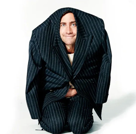Jake Gyllenhaal wearing an oversized pinstripe suit pulled over his head, kneeling against a white backdrop.
