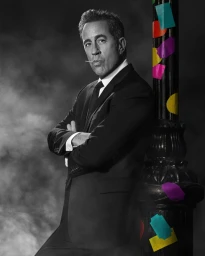 Jerry Seinfeld in a black suit with a lollipop in his mouth leaning against a lamppost with colorful stickers, photographed by Mary Ellen Matthews.