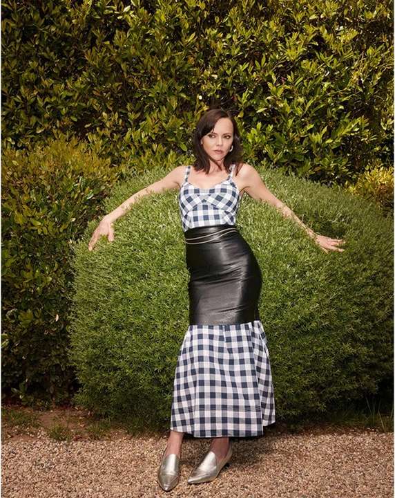 Adult female model poses in a black and white checkered top and black leather skirt in front of a green leafy background.