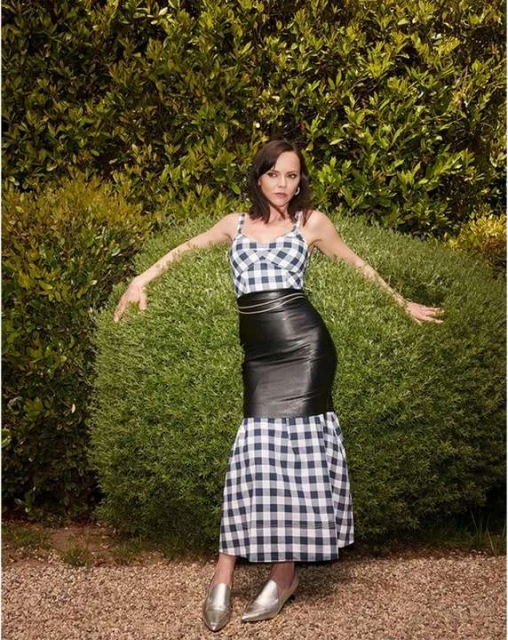 Adult female model poses in a black and white checkered top and black leather skirt in front of a green leafy background.
