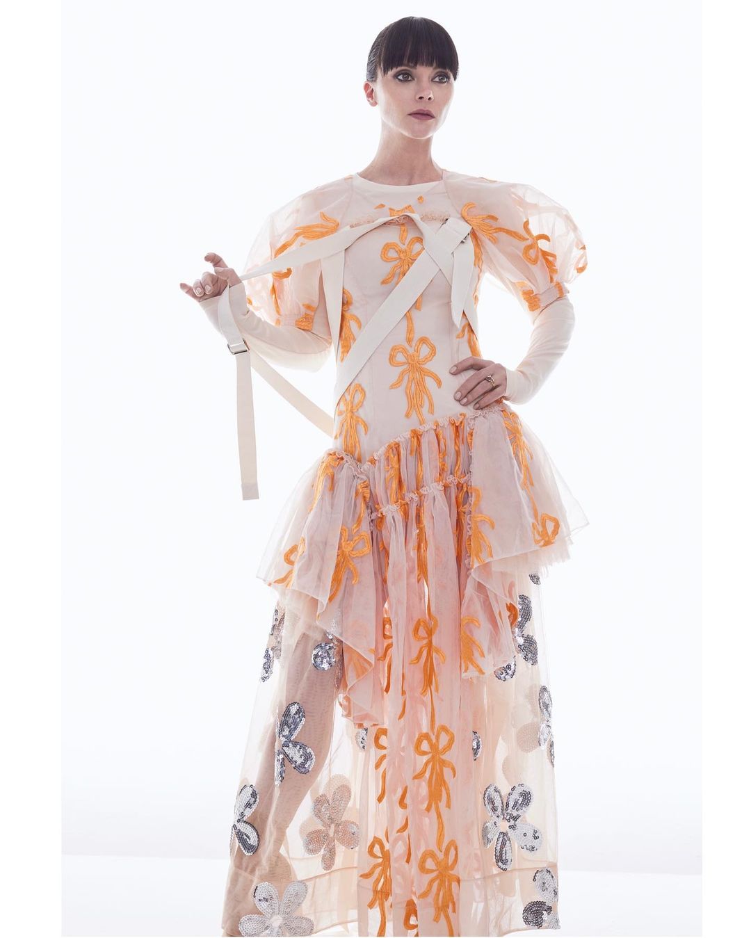 A woman in a light-peach couture gown poses against a bright white background. The gown is adorned with ribbon and flower details.