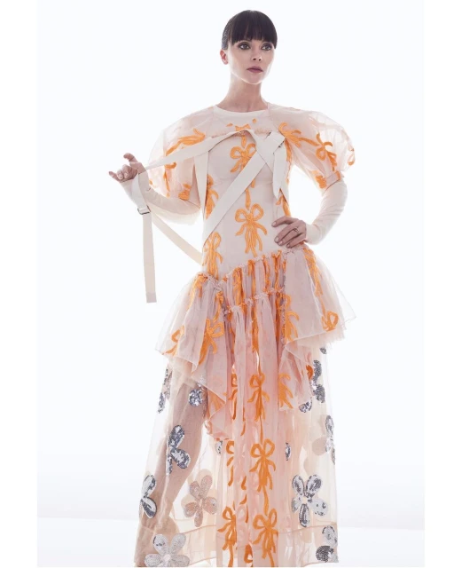A woman in a light-peach couture gown poses against a bright white background. The gown is adorned with ribbon and flower details.
