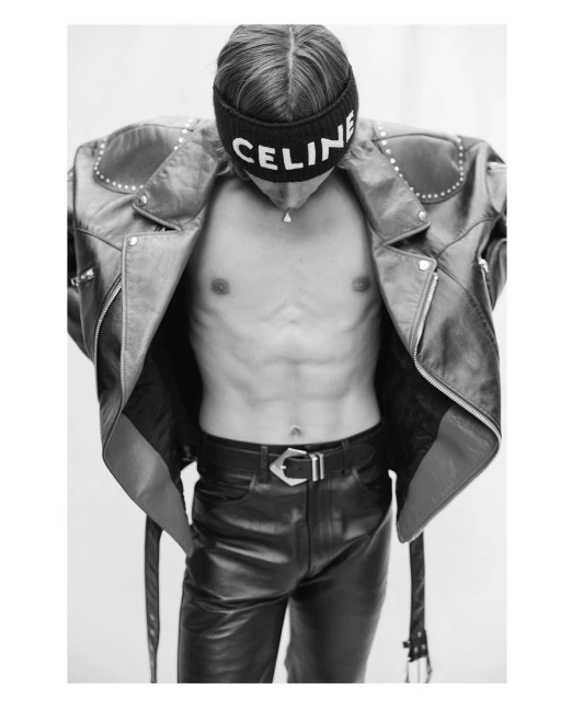 Black and white studio shot of a shirtless adult male model wearing a leather jacket, jeans, and beanie with a designer logo.