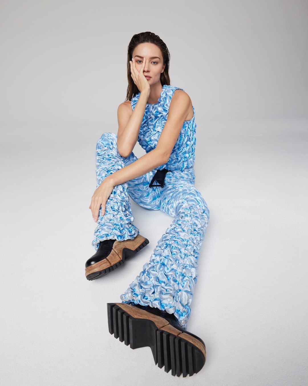 A stylish woman in a blue textured outfit poses confidently with chunky platform shoes against a neutral backdrop in a fashion shoot.
