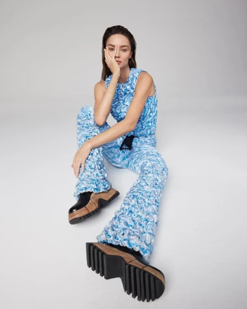 A stylish woman in a blue textured outfit poses confidently with chunky platform shoes against a neutral backdrop in a fashion shoot.