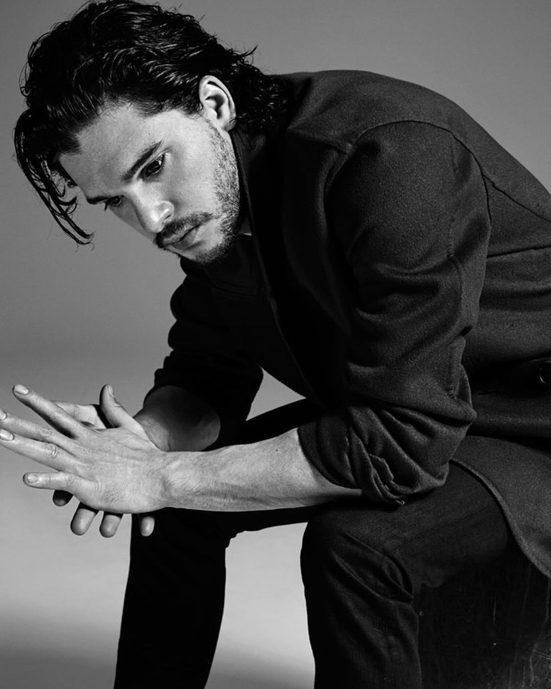 Portrait of an adult male with dark hair, wearing a black jacket, posing with his hands interlaced in a photo studio.