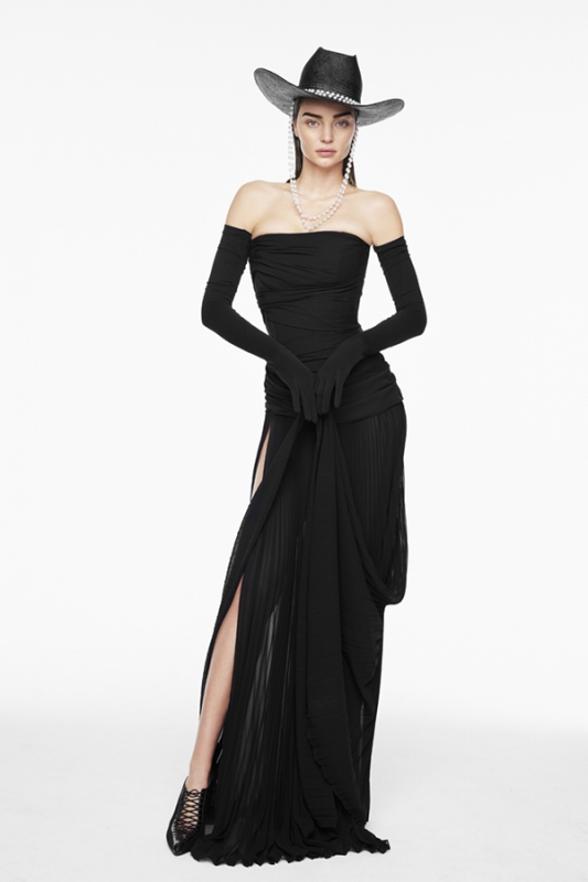 Woman in black strapless dress and cowboy hat stands in a studio, showcasing fashion-forward style and elegant design on a white backdrop.