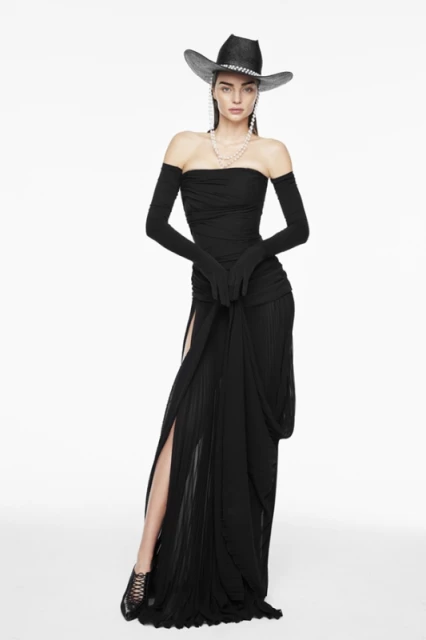 Woman in black strapless dress and cowboy hat stands in a studio, showcasing fashion-forward style and elegant design on a white backdrop.