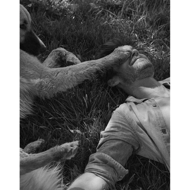 Monochrome image of a man lying on grass with a dog placing its paw over his eyes, creating a touching and intimate scene.