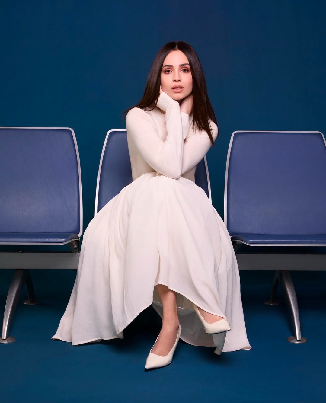 Sofia Carson seated on a blue chair wearing a long white dress and heels against a dark blue background