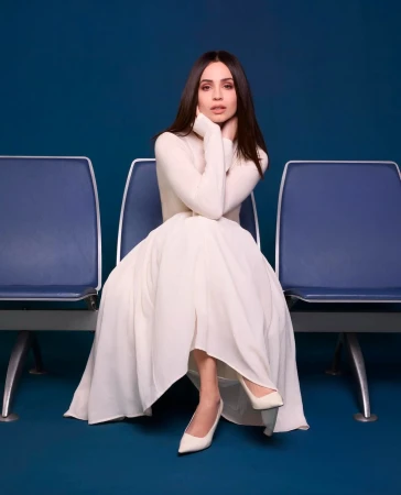 Sofia Carson seated on a blue chair wearing a long white dress and heels against a dark blue background