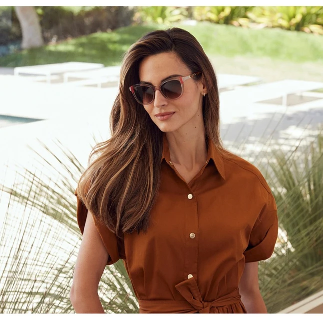 Brunette woman in a brown button-down dress and sunglasses, outdoors with greenery and light-colored hardscape in the background.
