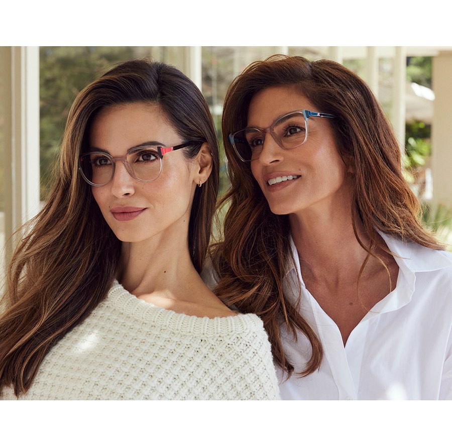 Two adult women pose indoors wearing eyeglasses and casual clothing, against a background of windows and greenery.