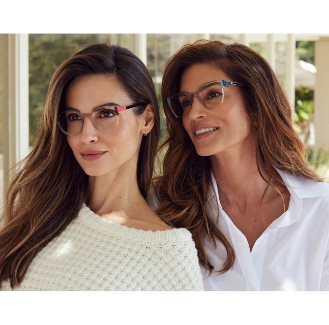 Two adult women pose indoors wearing eyeglasses and casual clothing, against a background of windows and greenery.
