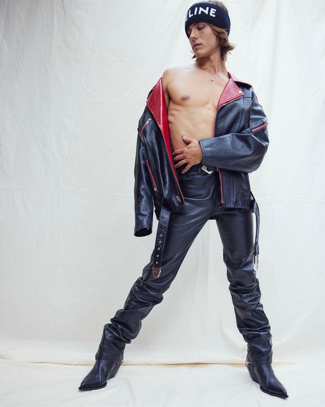 A slender man poses in a black leather jacket, black leather pants and black leather boots against a cream-colored backdrop.