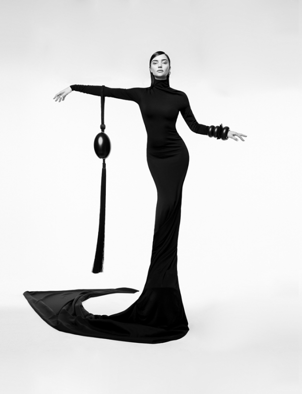 Model in a long black fitted dress with a sculptural tassel accessory photographed by Nino Munoz against a white backdrop.