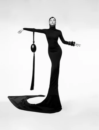 Model in a long black fitted dress with a sculptural tassel accessory photographed by Nino Munoz against a white backdrop.