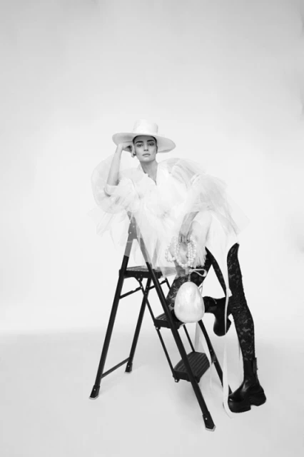 Adult woman sitting on a black ladder posing in a white hat and a white tulle dress in a photo studio with a pearl handbag.