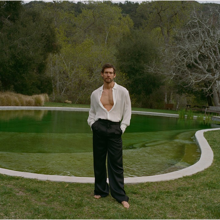 Male fashion model poses barefoot in a white unbuttoned shirt and black trousers near a green pool in an outdoor setting with trees.
