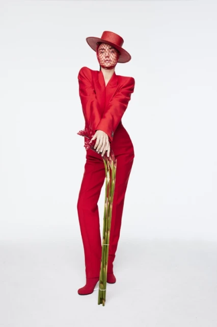 Woman poses in a red hat, veil, and pantsuit, holding a bundle of gladiolus flowers against a stark white backdrop.