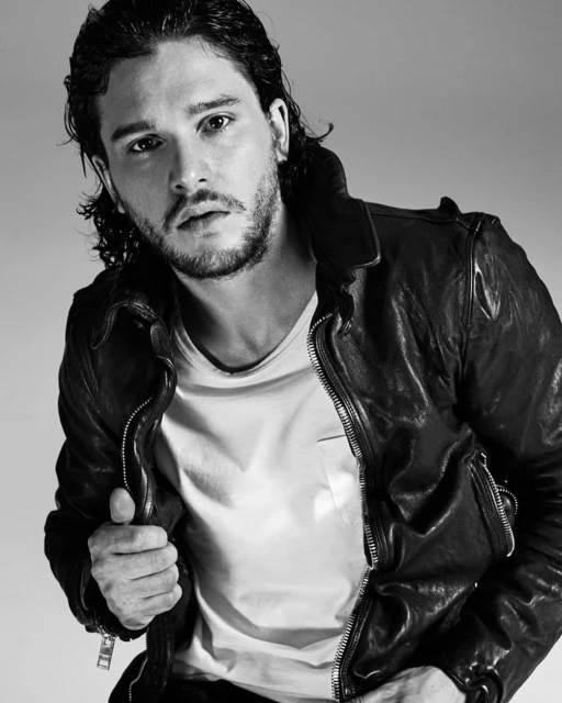 Black and white portrait of a man with dark, curly hair wearing a leather jacket and a white t-shirt in a studio setting.