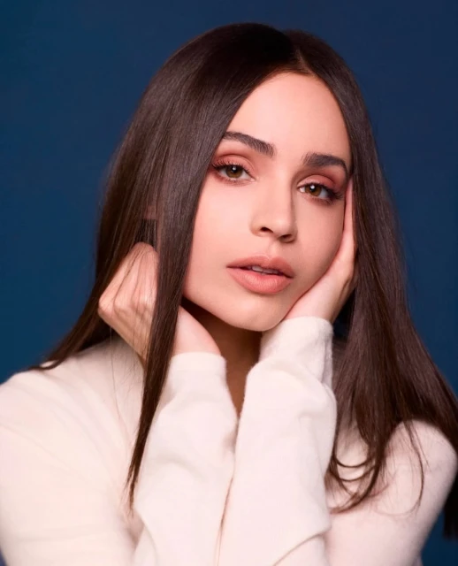 Portrait of a brunette woman with her hands on her face wearing a cream-colored sweater against a dark blue backdrop in eye-level shot.