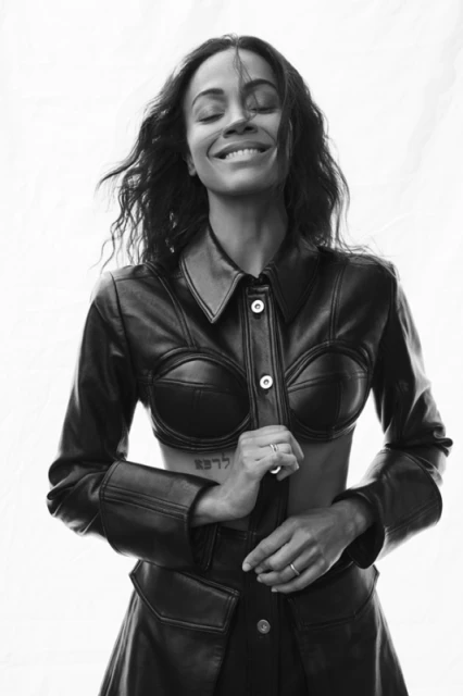 Black and white portrait of a smiling adult woman with dark hair, wearing a black leather outfit against a bright white background.