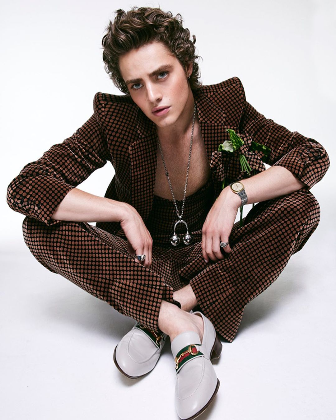 A young adult male model poses in a brown patterned suit with white loafers on a white backdrop, looking directly at the camera.