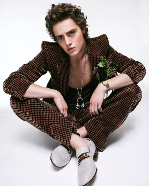 A young adult male model poses in a brown patterned suit with white loafers on a white backdrop, looking directly at the camera.