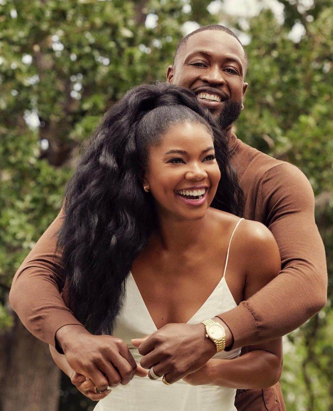 Smiling couple embraces outdoors: An African-American man hugs a woman with long, dark hair in a white dress against a green backdrop.
