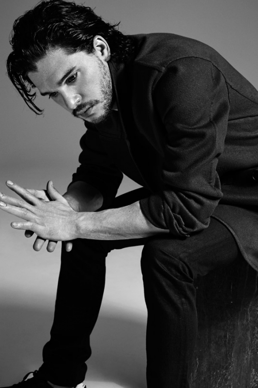 Monochrome portrait of a stylish adult male with dark hair, a light beard, and wearing a dark collared shirt and dark jeans while sitting.