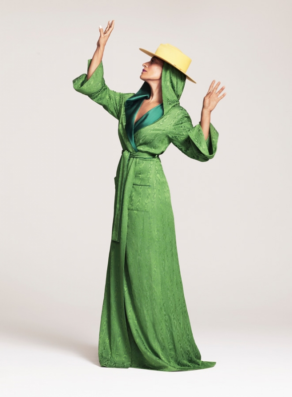 A woman in a full-length green robe and a yellow hat is posing with her hands up against a plain white background in a fashion shot.