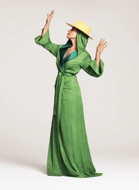 A woman in a full-length green robe and a yellow hat is posing with her hands up against a plain white background in a fashion shot.